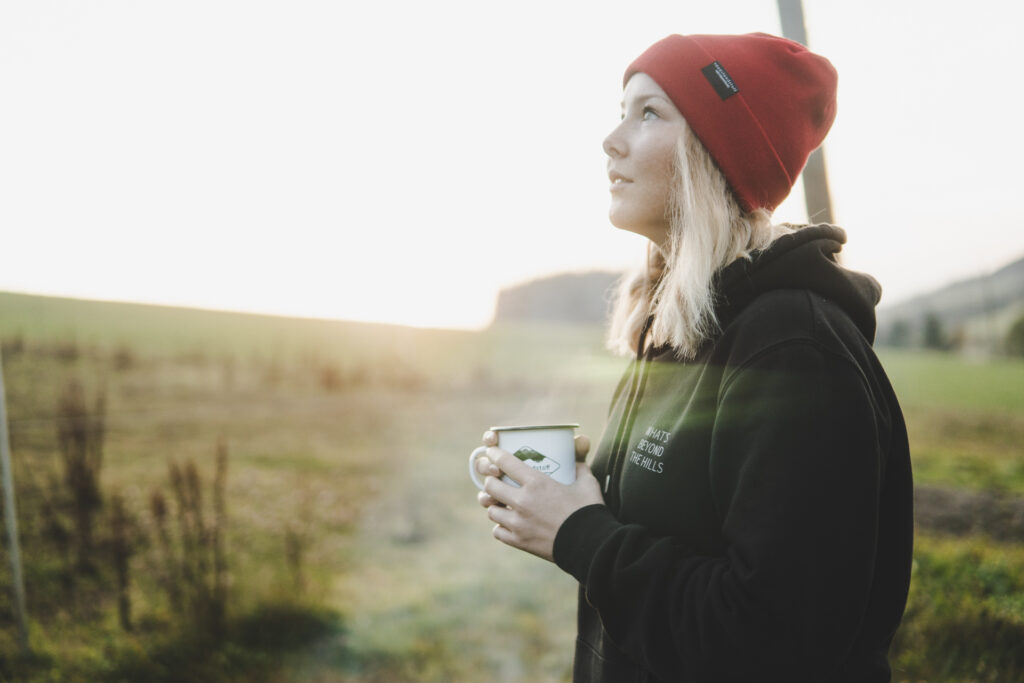 Eine junge Frau steht vor einem Lagerfeuer, hält eine Kaffetasse in der Hand und blickt in Richtung Himmel